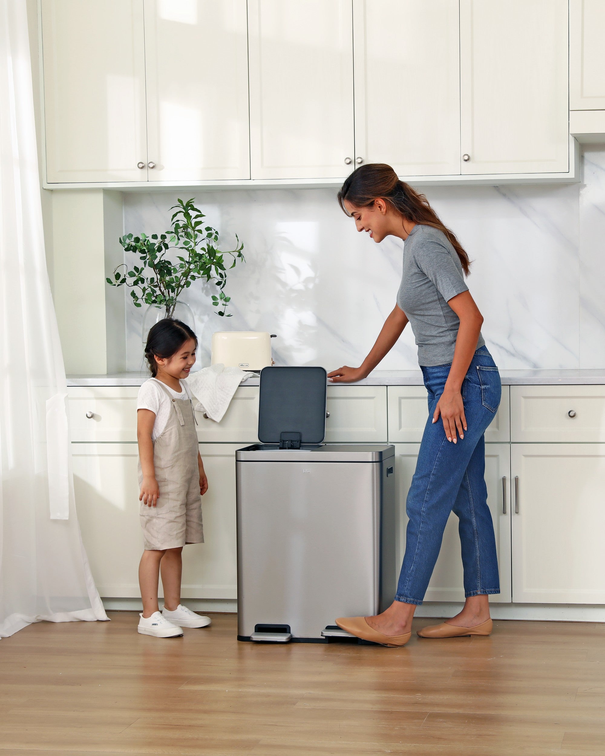 Woman and child in a kitchen with a trash can and recycling bin.